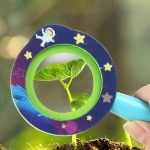 Nahaufnahme eines grünen Pflanzenblatts, sichtbar durch ein kindliches Lupenglas mit Weltraummotiv, gehalten von einer Hand. Perfekt für wissenschaftliche und Bildungszwecke.