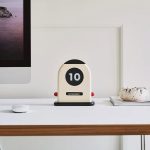 Moderne Kalenderuhr im Retro-Design auf einem weißen Schreibtisch, zeigt den 10. November, umgeben von einem Computerbildschirm und einer dekorativen Pflanze.