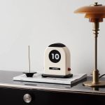 Stilvoller Retro-Wecker im Vintage-Design auf einem Schreibtisch, umgeben von Zeitschriften und einer modernen Tischlampe, ideal für Dekoration und funktionale Zeitmessung.