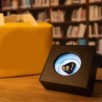 Modernes wissenschaftliches Gadget in schwarzem Gehäuse mit digitaler Anzeige, auf einem Holzschreibtisch vor Bücherregal, ideal für Wissenschafts- und Dekorationsliebhaber.
