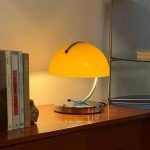 Helle gelbe Tischlampe mit gebogener Metallhalterung auf einem Holzschreibtisch, neben Büchern und Dekorationsobjekten, in einem stilvollen Raum.