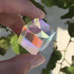 Hand hält einen transparenten Kristallwürfel mit Regenbogeneffekt vor grünen Blättern, der Licht bricht und bunte Reflexionen erzeugt.