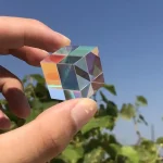 Hand hält einen bunten, reflektierenden Kristallwürfel vor blauem Himmel, umgeben von grünen Blättern, als wissenschaftliches Dekorationsstück.