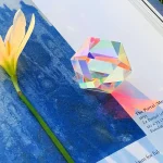 Sternförmiger Kristall mit holographischem Glanz auf blauer Oberfläche, neben einer weißen Blume. Perfekt für wissenschaftliche Dekorationen und Sammlerstücke.