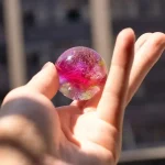 Hand hält einen bunten, glitzernden Stein mit rosa, goldenen und transparenten Elementen, vor einem unscharfen Hintergrund. Perfekt für wissenschaftliche Dekorationen und Sammlerstücke.