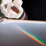Nahaufnahme eines Sonnenstrahls, der auf eine glatte Oberfläche trifft und einen Regenbogen-Effekt erzeugt, während eine Person mit einem weißen Handschuh einen prismatischen Gegenstand hält.