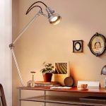 Moderne Schreibtischlampe mit verstellbarem Arm auf einem Holzschreibtisch, beleuchtet einen Arbeitsbereich mit Pflanzen, Büchern und Dekorationen.