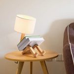 Kreatives Dekorationsstück eines kleinen Holz-Menschen, der auf einem Tisch sitzt, mit einem Buch in den Armen und einer Lampe im Hintergrund, ideal für moderne Wohnräume.