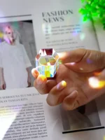Hand hält einen transparenten, facettierten Kristall, der Licht bricht und Regenbogenfarben reflektiert, vor einem Magazin mit Mode- und Wissenschaftsthemen.