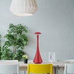 Elegante, rote Tischlampe mit schlankem Design auf einem weißen Tisch, umgeben von Pflanzen und moderner Dekoration, ideal für stilvolle Wohn- oder Büroräume.
