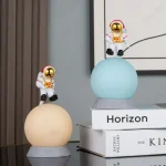 Zwei kleine Astronautenfiguren in Raumanzügen mit goldenen Helmen, die auf bunten, leuchtenden Planeten sitzen, ideal für wissenschaftliche Dekorationen und Raumfahrt-Themen.