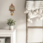 Detaillierte Ansicht einer dekorativen Schmuckuhr im Chalet-Stil an einer weißen Wand, neben einem weißen Tisch mit Topfpflanze, und einem Holzgestell mit weißen Pompons.