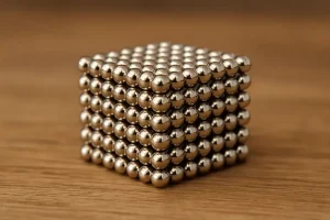 Ein 3D-Modell eines Würfels, bestehend aus kleinen, glänzenden Metallkugeln, die magnetisch verbunden sind, auf einem Holzuntergrund.