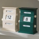 Zwei dekorative Flip-Uhren, eine in Creme und eine in Dunkelgrün, zeigen das Datum und den Wochentag, ideal für stilvolle Raumdekorationen und funktionale Kalender.