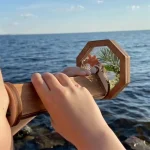 Hand hält ein Holzobjekt mit Spiegel und kleinen Pflanzen vor dem Meer, ideal für wissenschaftliche Dekorationen und kreative Raumgestaltung.