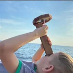 Kind hält ein Teleskop-Periskop am Meer, blickt durch es und entdeckt die Weite des Ozeans bei Sonnenuntergang. Perfekt für Wissenschafts- und Entdeckungsfans.