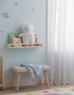 Weiches Weltraum-Spielzeug in einem hellblauen Kinderzimmer, mit Bücherregal, Stern- und Planeten-Wanddekoration, auf einem kleinen Sitzhocker mit Decke.