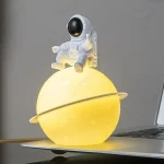 Niedliche Raumfahrer-Figur auf einer leuchtenden Planet-Lampe, ideal als Dekoration für Wissenschafts- und Raumfahrtliebhaber.