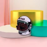 Dekorativer Raumfahrt-Button mit sowjetischem Bären in Astronautenhelm, auf bunten geometrischen Plattformen präsentiert.