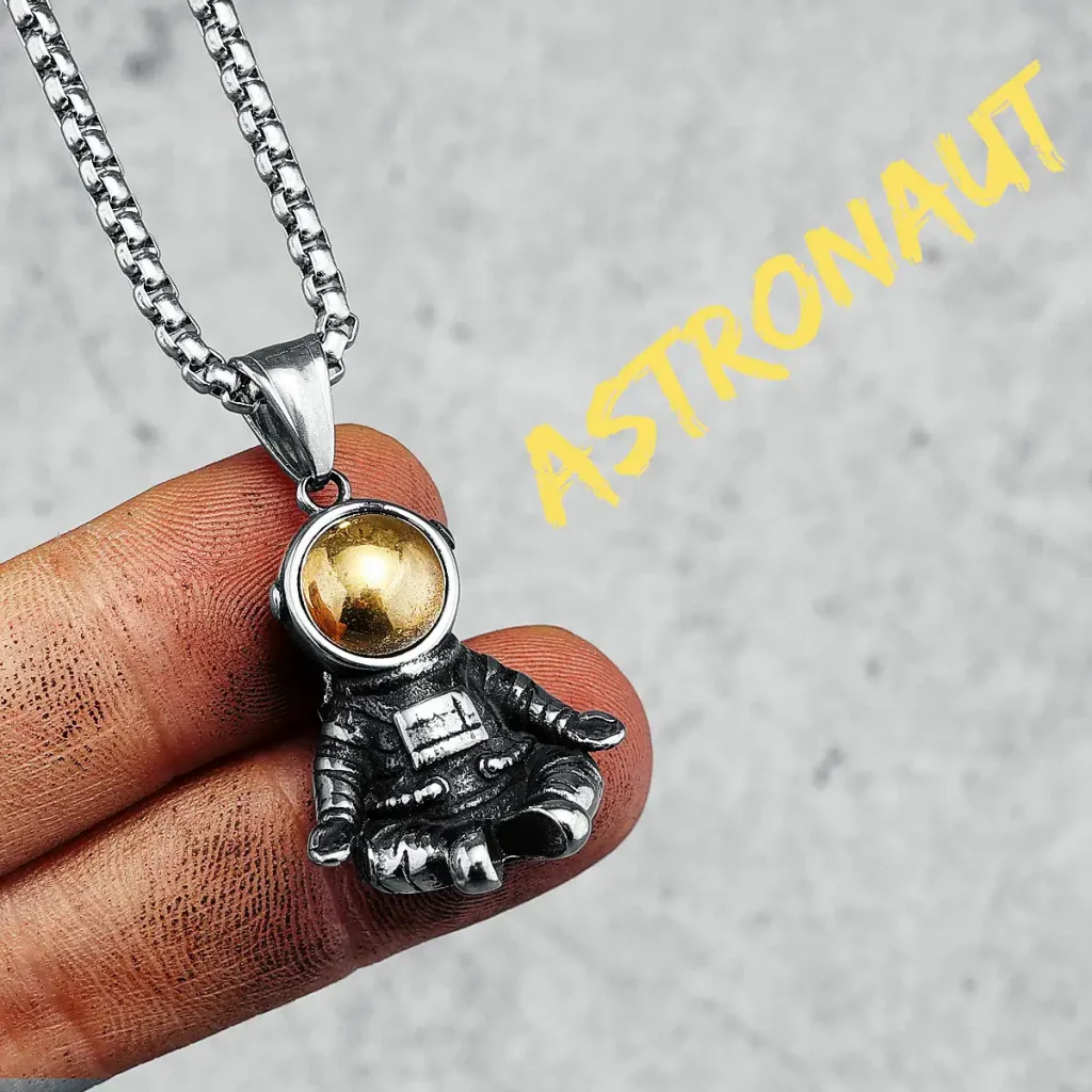 Detailaufnahme eines Astronauten-Anhängers mit goldener Helmkapsel, gehalten zwischen den Fingern, auf grauem Hintergrund.