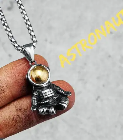 Detailaufnahme eines Astronauten-Anhängers mit goldener Helmkapsel, gehalten zwischen den Fingern, auf grauem Hintergrund.