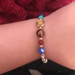 Handgelenk mit buntem Perlenarmband aus Himbeer- und Türkisfarbenen Perlen, kombiniert mit goldenen Akzenten, auf weichem roten Stoff.