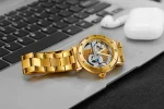 Hochwertige goldene Automatikuhr mit sichtbarem Uhrwerk, liegt auf einer Laptop-Tastatur, neben kabellosen Kopfhörern. Perfekt für Uhrenliebhaber und Technikfans.