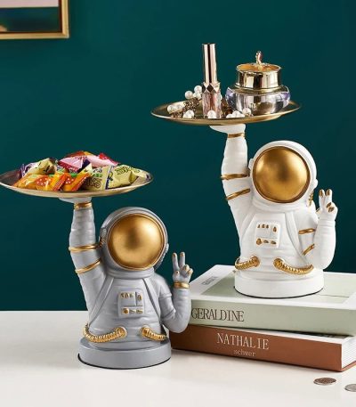 Zwei Astronauten-Dekorationsfiguren mit goldenen Helmen, die auf einer weißen Oberfläche stehen, halten silberne Tabletts mit Schmuck und Süßigkeiten, ideal für Science-Fiction-Dekor.