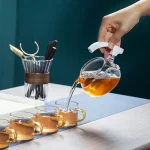 Person gießt Tee aus einem wissenschaftlichen Glasgefäß in drei Tassen, auf einem weißen Tisch mit Schreibutensilien im Hintergrund.