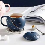 Dekorative Tasse im Weltraumdesign mit passendem Löffel und Maus, ideal für Kaffee oder Tee, auf einem weißen Tisch mit Buch im Hintergrund.