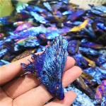 Bunt schillernder, blauer Kristall mit metallischem Glanz, gehalten in der Hand vor einem Hintergrund aus weiteren ähnlichen Kristallen.
