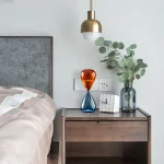 Modernes Schlafzimmer mit Nachttisch, dekorativem Glasobjekt in Orange und Blau, sowie einer Vase mit Eukalyptus, beleuchtet durch eine elegante Deckenlampe.