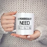 Weiße Tasse mit wissenschaftlichem Text "I Periodically NEON" und chemischen Elementen, gehalten von einer Person in einem grauen Pullover.