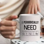 Weiße Tasse mit dem Spruch "I PERIODICALLY NEED" und einem Periodensystem-Design, das wichtige chemische Elemente zeigt. Perfekt für Wissenschafts- und Chemie-Liebhaber.