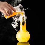 Wissenschaftliches Chemie-Experiment mit Reagenzglas, das eine orange Flüssigkeit enthält, und einer Flasche, die Rauch oder Dampf freisetzt, auf schwarzem Hintergrund.