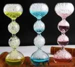 Drei bunte Glas-Objekte mit Blasenmuster, die auf einem weißen Tisch stehen, jeweils in Gelb, Blau und Rosa, vor einem schwarzen Hintergrund.