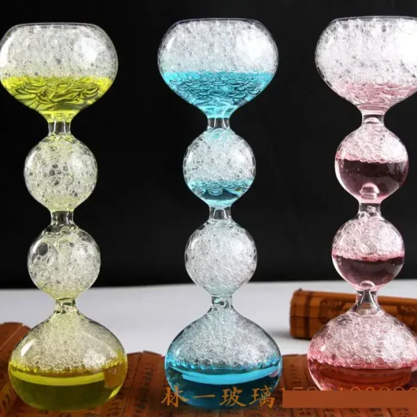 Drei bunte Glas-Objekte mit Blasenmuster, die auf einem weißen Tisch stehen, jeweils in Gelb, Blau und Rosa, vor einem schwarzen Hintergrund.