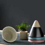Verschiedene geometrische Formen aus Keramik und Holz, inklusive Pyramiden, Würfel und Scheiben, auf einer schwarzen Oberfläche mit grünem Pflanzen im Hintergrund.