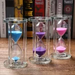 Sanduhr mit bunten Sandkörnern in drei Farben: Blau, Lila und Pink, in transparenten Glasgehäusen, auf einem Holzregal mit Büchern im Hintergrund.
