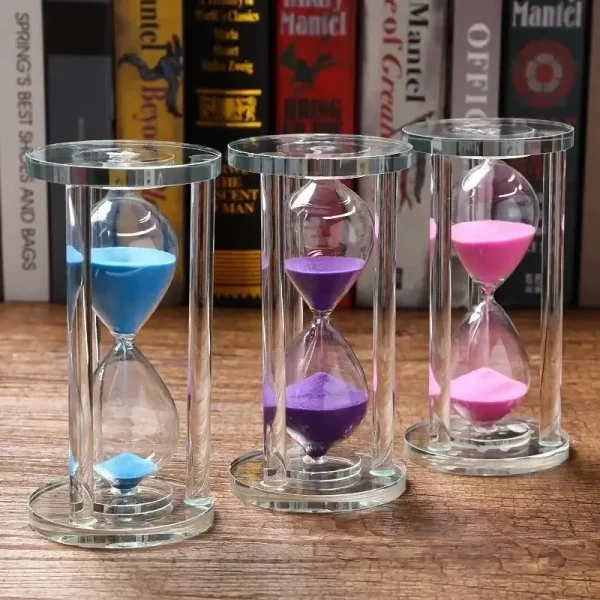 Sanduhr mit bunten Sandkörnern in drei Farben: Blau, Lila und Pink, in transparenten Glasgehäusen, auf einem Holzregal mit Büchern im Hintergrund.