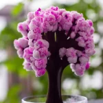 Nahaufnahme einer einzigartigen, blumenähnlichen Kristallformation mit rosa und weißen Farbnuancen, die auf einem dunklen Stiel wächst, vor einem unscharfen grünen Hintergrund.