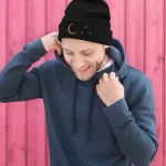 Junge trägt eine schwarze Mütze mit Mond- und Sternmotiven vor einem pinken Holzzaun.