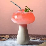 Kreatives Blumenmushroom-Getränk mit rosa Flüssigkeit, Minzblatt und Strohhalm, auf einem dekorativen Untergrund mit Blättern und Salz.