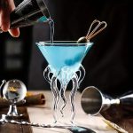 Ein wissenschaftliches Cocktail-Glas mit blauer Flüssigkeit, das auf einem Tisch steht, mit einem Hand, die die Flüssigkeit einschenkt, und einem dekorativen Tentakel-Design.