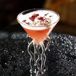 Ein stilvolles wissenschaftliches Cocktailglas mit einem kunstvollen, gewundenen Glasständer, gefüllt mit einem rosa Getränk, garniert mit Blütenblättern.