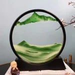 Kunstvolles Sandbild in einem schwarzen Kreisrahmen, das eine malerische Berg- und Landschaftsszene mit grünen Sandstrukturen zeigt.
