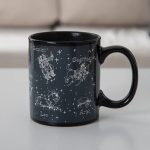 Schwarze Tasse mit weißen Sternzeichen-Grafiken der Tierkreiszeichen, darunter Zwillinge, Löwe, Schütze und Widder, auf einem weißen Hintergrund.