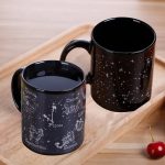Zwei schwarze Tassen mit Sternzeichen-Designs, die auf einem Holztablett liegen, mit einem Kirschen im Vordergrund.