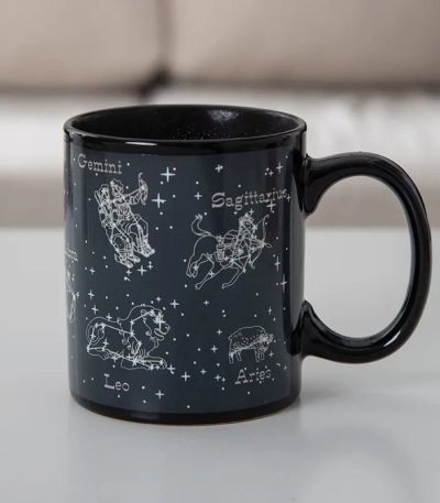 Schwarze Tasse mit weißen Sternzeichen-Grafiken der Tierkreiszeichen, darunter Zwillinge, Löwe, Schütze und Widder, auf einem weißen Hintergrund.