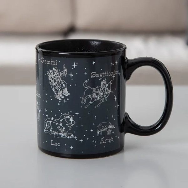 Schwarze Tasse mit weißen Sternzeichen-Grafiken der Tierkreiszeichen, darunter Zwillinge, Löwe, Schütze und Widder, auf einem weißen Hintergrund.